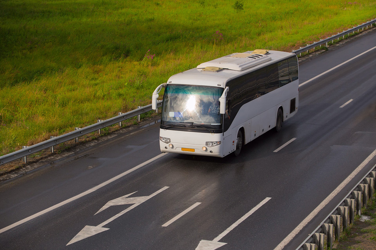 A white motorcoach-style bus drives along a well paved road, passing through a lush green field.